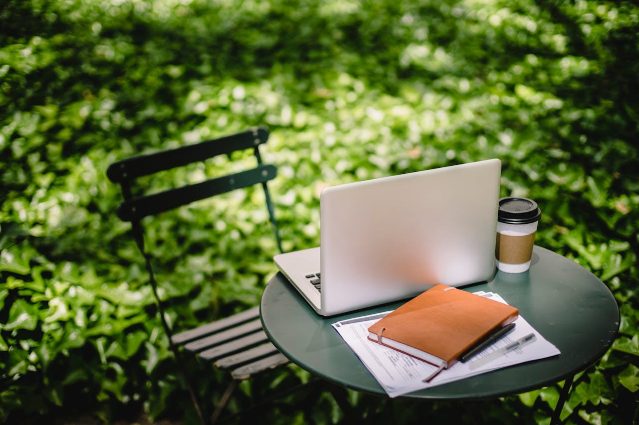 Modern netbook with notebook placed on small round table near paper cup with hot drink in sunny green park